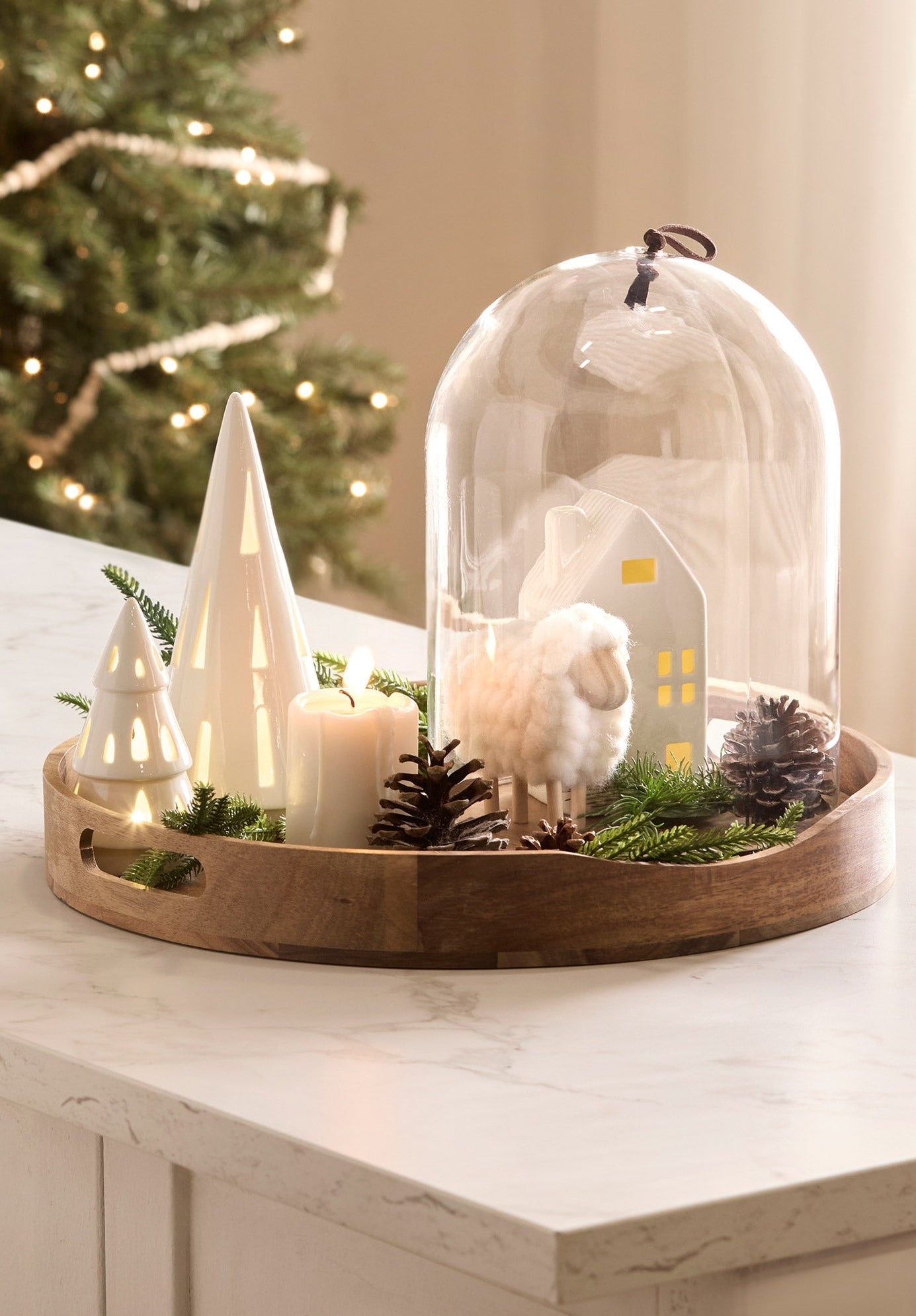 Decorative setup with a wooden tray containing a sheep figurine, candles, and Christmas trees on a marble surface.